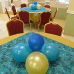 (Balloon Table Setting)
​Continuing with the simple, elegant and festive decor, the foundation of the 12 tables were created with gold synthetic table coverings. A teal with gold metallic speckled fabric was hand cut and used as the overlay. ~