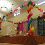 (Paper Garland Mandap)
This mandap was created using bright multi colored paper garlands wrapped around the upper horizontal and vertical perimeters. Several yards of solid and printed fabrics made a more interesting backdrop than the plain wall. Traditional mango leaves topped the pillars. ~