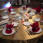 (Round Table Design and Settings)
​Gold satin cloths blanketed each table. A sheer ivory scroll fabric was used as the runner. Red cloth napkins were gathered into a "purse" design with a rhinestone napkin ring. The center of the table held a low, clear glass bowl and within floated a single white or pink lotus flower. Peacock feathers, mini red "jems" and faux rose petals were among the adornments that completed this beautiful setting. ~