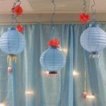 (Hanging & Backdrop Decor for Food Table)
Instead of seeing a plain, ugly wall behind the food table, blue fabric was hung as a backdrop. White lights twinkled from underneath while fresh lotus flowers dangled amongst. A trio of blue paper lanterns adorned with beading hung directly over the the table as a decorative focal point. Glitter "squiggles" and coral colored silk lotus flowers added to this simple but embellishing display. (Lanterns with beading available as a rental for your function) ~